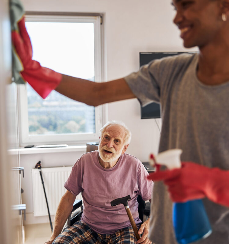 Aged man in the wheelchair watching a smiling young African American caregiver polishing his wardrobe
